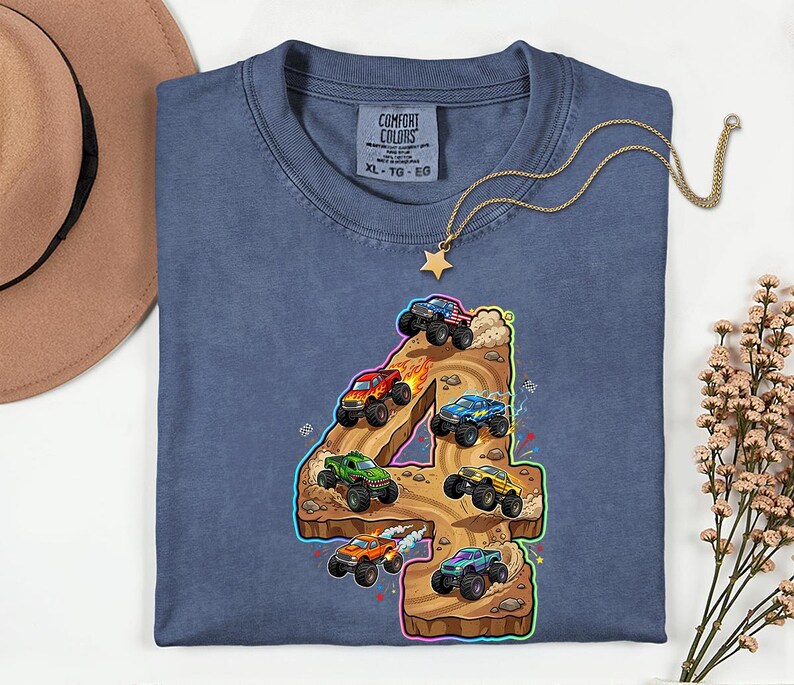 May include: A blue Comfort Colors t-shirt with a vibrant graphic of monster trucks on a number 4. The design showcases monster trucks in red, green, blue, and yellow, with flame and dust effects. A gold necklace with a star pendant is on the shirt.