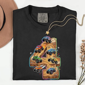 May include: Black crew neck sweatshirt featuring a colourful graphic of monster trucks racing on a number 4. The graphic includes red, green, blue, and yellow trucks. A gold necklace with a star pendant is on the shirt.
