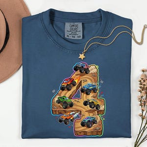 May include: A navy blue t-shirt featuring a colourful graphic of monster trucks racing on a number 4. The graphic includes various monster trucks in red, green, blue, and orange. The shirt has a crew neck and a tag that says "Comfort Colors".