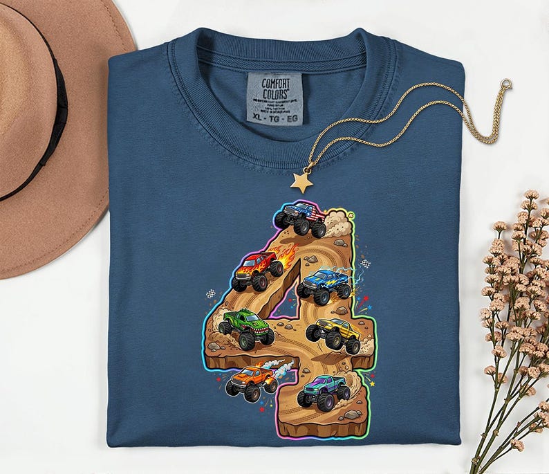 May include: A navy blue t-shirt featuring a colourful graphic of monster trucks racing on a number 4. The graphic includes various monster trucks in red, green, blue, and orange. The shirt has a crew neck and a tag that says "Comfort Colors".