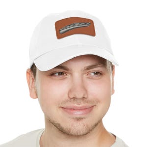 May include: White baseball cap with a brown leather patch featuring a silver canoe design. The cap has a curved brim and an adjustable strap. This hat is a stylish accessory for outdoor enthusiasts.