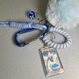 May include: A collection of blue accessories, including a carabiner, a keychain with a Dragonair card, a fuzzy pom-pom, a coiled keychain, and a blue dice. The items are arranged on a light gray surface.