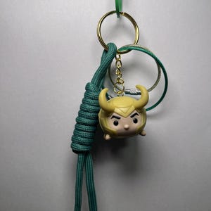 May include: A Loki Tsum Tsum keychain with a teal lanyard and gold-colored accents. The keychain features a small, round Loki figure with a yellow helmet and horns. The lanyard is attached to a green clip and a gold keyring.