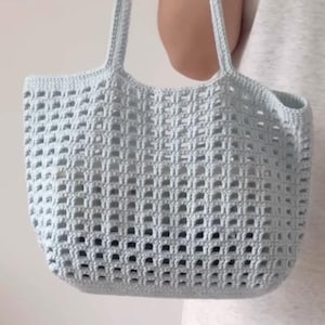 Könnte beinhalten: Hellblaue Häkeltasche mit zwei Henkeln. Die Tasche hat ein quadratisches, offenes Webmuster. Die Tasche wird von einer Person gehalten.