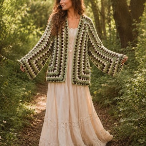 Op de afbeelding: Een gehaakt vest met lange mouwen en een granny square patroon in tinten donkergroen, lichtgroen en bruin. Het vest heeft een open voorzijde en wijde mouwen. Het wordt gedragen over een enkellange, crèmekleurige jurk met kanten details.