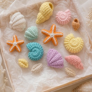 Può includere: Una collezione di figure di vita marina all'uncinetto che include due stelle marine arancioni con centri bianchi, varie conchiglie a spirale e a ventaglio in tonalità pastello di giallo, rosa, azzurro, lavanda, bianco e beige, disposte all'interno di una scatola.