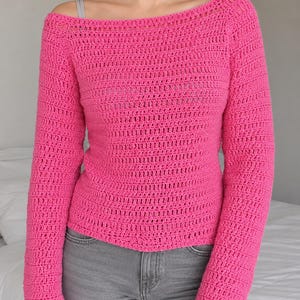 May include: A bright pink, long-sleeved crochet sweater with a boat neck. The sweater is made of a textured yarn and has a fitted silhouette. The model is wearing gray jeans.