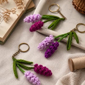 May include: Handmade crochet keychains featuring lavender flower designs. Each keychain has a gold-colored ring and chain, with green leaves and two lavender-colored flower heads in shades of purple. The keychains are displayed on a neutral-colored surface.