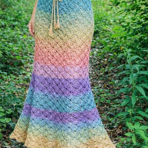 May include: A long, handmade crochet skirt in a rainbow of colours, including blue, green, yellow, orange, and purple. The skirt has a drawstring waist and a flared hem. The skirt is worn with a white top and brown sandals.