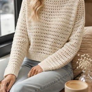 Puede incluir: Suéter de punto crema con mangas largas y cuello barco. El suéter presenta un patrón texturizado con detalles acanalados horizontales en las mangas y el cuerpo. La persona lleva vaqueros azul claro. Un estilo casual y cómodo.