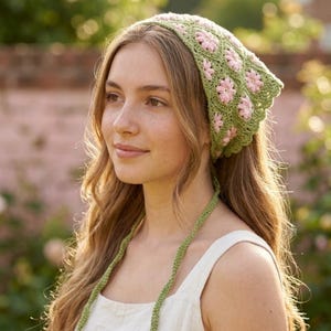 Puede incluir: Un pañuelo verde tejido a ganchillo con flores rosas. El cubrecabezas está atado en la parte posterior y tiene un borde festoneado. El pañuelo está hecho de un hilo suave y texturizado. La persona tiene el pelo largo y ondulado.