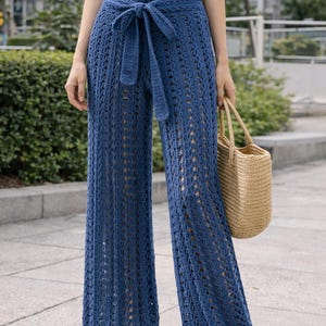 May include: Blue crochet palazzo trousers with a matching tie belt. The trousers have a wide leg design and are paired with a white top and a woven tote bag. The trousers are made of a breathable, open-weave fabric.