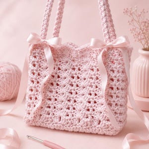 May include: A light pink crocheted tote bag with two braided handles and satin ribbon bows. The bag features an openwork design and is displayed with a crochet hook, a ball of yarn, and a vase with flowers. The overall theme is handmade crafts.