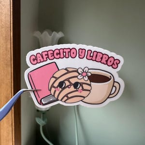 Op de afbeelding: Een sticker met de tekst "Cafecito y Libros" in roze letters. De sticker toont een roze boek, een concha-gebakje met een bloem en een koffiekopje. De sticker wordt vastgehouden door een pincet.