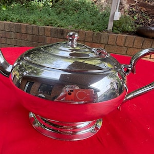 May include: A polished silver-tone metal teapot featuring a rounded body, a curved spout, a matching lid with a small knob handle, and a decorative arched handle. The reflective surface shows a distorted view of the surrounding environment.