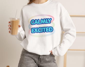 May include: A white long-sleeve sweatshirt featuring the text CALMLY EXCITED in blue bubble letters. The text is centered on the chest and surrounded by three faint, blurred horizontal oval rings in pastel pink, blue, and green colors.