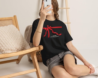 May include: A black short-sleeved t-shirt featuring a graphic of a bright red ribbon tied in a bow across the chest. Below the ribbon, the text reads CAUTION UNWRAP CAREFULLY AND ENJOY. The shirt has a crew neckline and a relaxed, casual fit.