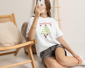 May include: A white short-sleeve t-shirt featuring a graphic of a Christmas tree made of green tennis balls, topped with a small green creature, and a tennis racket at the base. The text Merry Christmas is printed in red above the tree design.