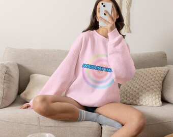 May include: A light pink long-sleeve crewneck sweatshirt featuring a central graphic of a multicolored pastel spiral. The text "ANXIOUSLY HAPPY" is printed in a bold blue font across the center of the spiral design on the front of the garment.