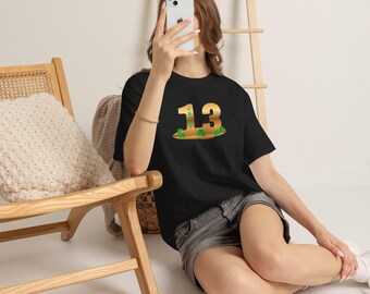 May include: A black short-sleeved t-shirt featuring a large gold-colored graphic of the number 13 with the word LUCKY written vertically inside the number one, accented by small green shamrock illustrations at the base of the digits.