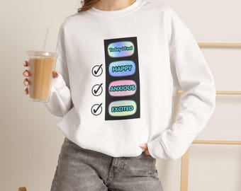 May include: A white long-sleeve sweatshirt featuring a vertical graphic with the text Today I Feel followed by three rounded rectangular buttons labeled Happy, Anxious, and Excited, each with a black checkmark icon to the left of the buttons.