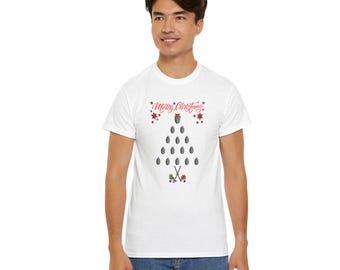 May include: A white short-sleeve t-shirt featuring a graphic of a Christmas tree made from grey hockey pucks. The design includes the red text Merry Christmas, red star accents, two crossed hockey sticks, and a pair of ice skates at the base.