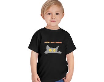 May include: A black short-sleeved t-shirt featuring a graphic of a grey cat with large yellow eyes peeking over the edge. Above the cat, the text HAPPY HALLOWEEN is printed in orange and yellow letters. The shirt has a crew neckline.