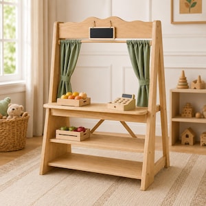 Könnte beinhalten: Ein Marktstand aus hellem Holz mit zwei salbeigrünen Stoffvorhängen, einer kleinen rechteckigen Kreidetafel oben, einer Spielzeugkasse aus Holz und zwei Holzkisten mit verschiedenen Spielzeugfrüchten auf den Regalböden.