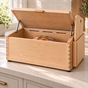 Puede incluir: Una panera de madera de tono claro con una tapa abatible sujeta por soportes metálicos. La caja presenta esquinas con juntas de caja, ranuras de ventilación en la parte posterior y un compartimento lateral que contiene una tabla de cortar.