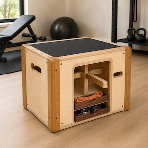 May include: A light-colored wooden plyometric box with a black textured top surface, rounded corner supports, and side cut-out handles. The interior features a cross-brace structure, a storage shelf with an orange resistance band, and a lower tray.