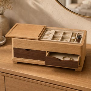 May include: A rectangular wooden jewelry box featuring a light oak exterior with two dark walnut-toned drawers. The top lid slides open to reveal a beige velvet-lined interior with multiple compartments for rings, watches, and small accessories.