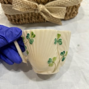 May include: Cream-colored teacup with green shamrock designs. The cup has a scalloped rim and a small handle. A woven basket with a bow is in the background.