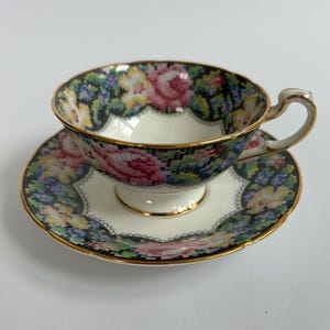 May include: A vintage teacup and saucer set. The cup and saucer are decorated with a mosaic floral pattern in pink, yellow, blue, and black. The rims are trimmed with gold. The cup has a curved handle.