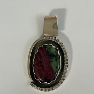 May include: A silver pendant with an oval-shaped ruby zoisite stone. The stone is a mix of deep red and green hues, set within a decorative silver frame. The pendant has a rectangular bail for a chain.