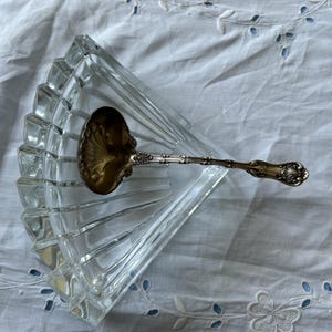 May include: A clear, fan-shaped glass dish with a decorative silver spoon resting inside. The spoon has an ornate handle and a gold-colored bowl. The dish and spoon are on a white, embroidered surface.