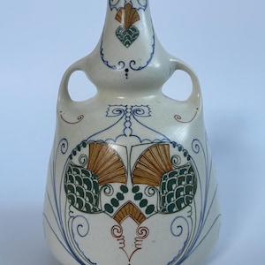 May include: A cream-colored ceramic vase with two handles and a narrow neck. The surface features intricate hand-painted designs in blue, green, and gold, including fan-like motifs, swirling lines, and geometric patterns arranged symmetrically.