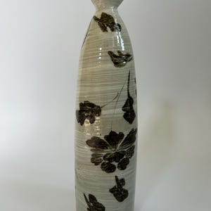 May include: A tall, ceramic vase with a narrow neck and a wide base. The vase is off-white with a textured, striped pattern and dark brown floral designs. The vase is approximately 12 inches tall.