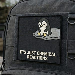 May include: Black backpack with a rectangular patch featuring a cartoon penguin on an ice floe. The patch reads "IT'S JUST CHEMICAL REACTIONS" in white text. The backpack has multiple straps and metal D-rings.