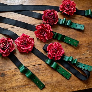 May include: Several black elastic bands with green leather accents and vibrant red rose embellishments are arranged on a wooden surface. The roses have detailed petals, and the green leather has a bow-like design. The bands appear to be accessories.