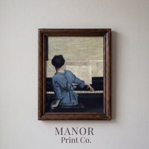 May include: Framed vintage print of a person playing a piano, wearing a blue dress. The artwork has a dark wooden frame and is mounted on a white wall. The text "MANOR Print Co." is printed at the bottom of the frame.