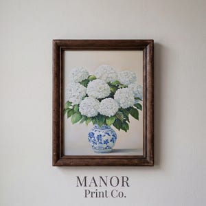 Pode incluir: Uma impressão emoldurada com uma natureza morta de hortênsias brancas num vaso azul e branco. A obra de arte está sobre um fundo neutro e emoldurada numa moldura de madeira castanha escura. O texto "MANOR Print Co." está na parte inferior.