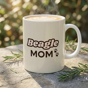 Puede incluir: Una taza de cerámica blanca con las palabras "Beagle MOM" en una fuente lúdica, acompañada de huellas de patas. La taza contiene una bebida caliente con un diseño decorativo de espuma. La taza está al aire libre.