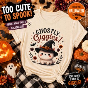 Puede incluir: Una camiseta de color crema con un gráfico de un gatito blanco y esponjoso que lleva un sombrero de bruja negro. El texto sobre el gatito dice "GHOSTLY Giggles!" en una fuente de color marrón oscuro. El diseño está rodeado de murciélagos.