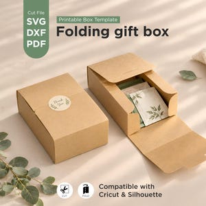 Pode incluir: Duas caixas de presente dobráveis de papel kraft castanho de formato retangular. Uma caixa está fechada com um autocolante redondo Thank You, enquanto a outra está aberta, revelando um pequeno pacote branco com uma ilustração de folhas verdes.