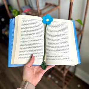 May include: A crochet bookmark featuring a bright blue flower with a yellow center attached to a long, dark green stem with a single leaf at the bottom, resting vertically against the open pages of a printed book with black text.