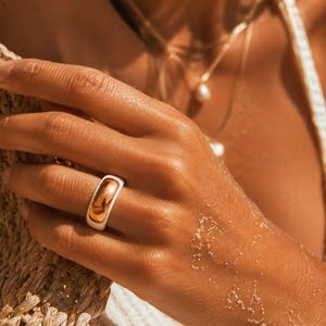 May include: A gold and white ring is worn on a hand. The ring has a rounded, smooth design with a gold interior and a white exterior. The hand is resting on a woven surface, and a necklace with pearls is visible.