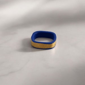 May include: A square-shaped ring with a vibrant blue exterior and a textured gold band. The ring is a bold statement piece, showcasing a modern design with contrasting colors and textures. This unique ring is a stylish accessory.