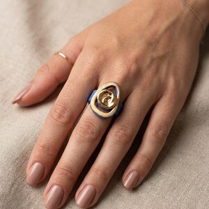May include: A gold ring with a blue base and a swirling design. The ring is on a hand with a matching gold band ring and a delicate bracelet. The nails are painted a neutral colour.