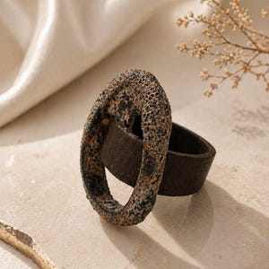 May include: A unique ring featuring a textured, oval-shaped top in shades of brown and black, set on a dark brown band. The ring's design has a rustic, weathered appearance, suggesting a vintage or handcrafted style. The ring is displayed on a neutral surface.