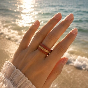 May include: A stack of three rings worn on a finger. The outer two rings are a solid terracotta red color with a smooth finish, while the center ring is a textured gold-toned band. The rings are worn together as a single set on one finger.